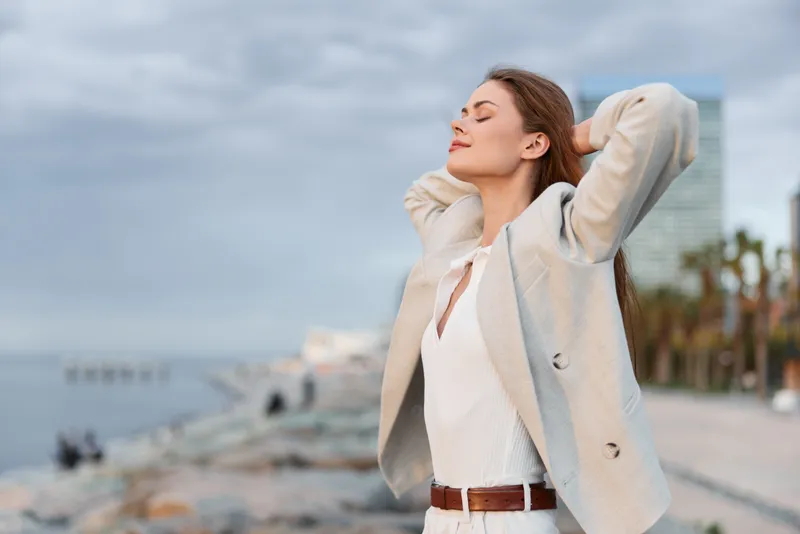 woman enjoying fresh air outdoors near waterfront, casual beige jacket and white outfit, serene expression, urban nature background, spring season, lifestyle concept