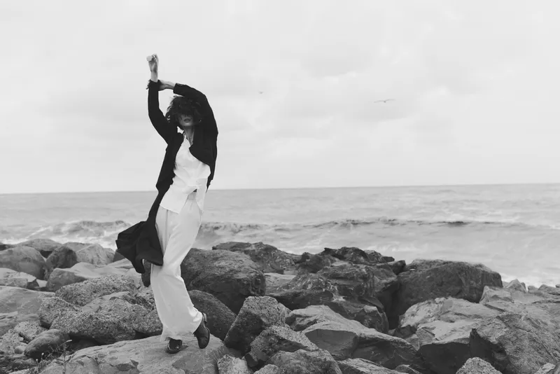 Dancer on a rocky shore by the sea, captured in motion with flowing white dress and dark jacket, dynamic pose against a vast seascape and cloudy sky