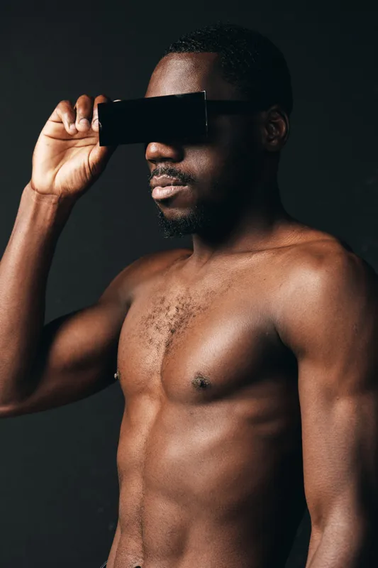 African man shirtless with dark skin holding black card in front of eyes in creative conceptual visual style on dark background. Male model posing with strong body and serious expression.