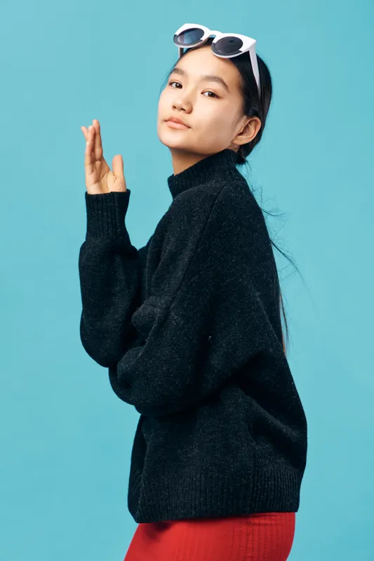 Young woman wearing black sweater and white glasses posing confidently against blue background with natural makeup and relaxed expression for fashion and style portrait.