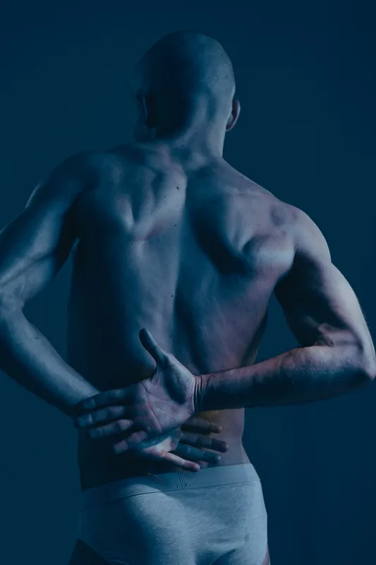 Male model with back pain, captured in a dramatic light, showcasing a strong physique with a blue hue, emphasizing discomfort and resilience in a striking composition.