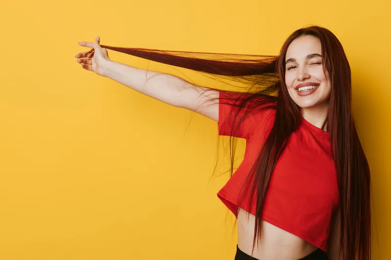 Young woman with long hair wearing red crop top smiling and pulling hair sideways against yellow background. Happy female posing with closed eyes and joyful expression in studio.