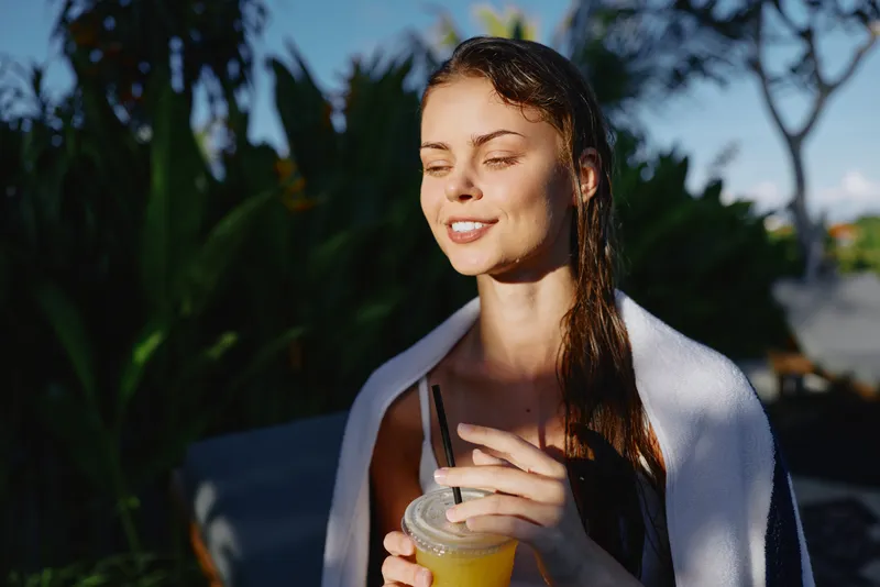 Young woman relaxes outdoors, wrapped in a towel and enjoying a cold drink with eyes closed, a peaceful sunlit moment that conveys a carefree lifestyle
