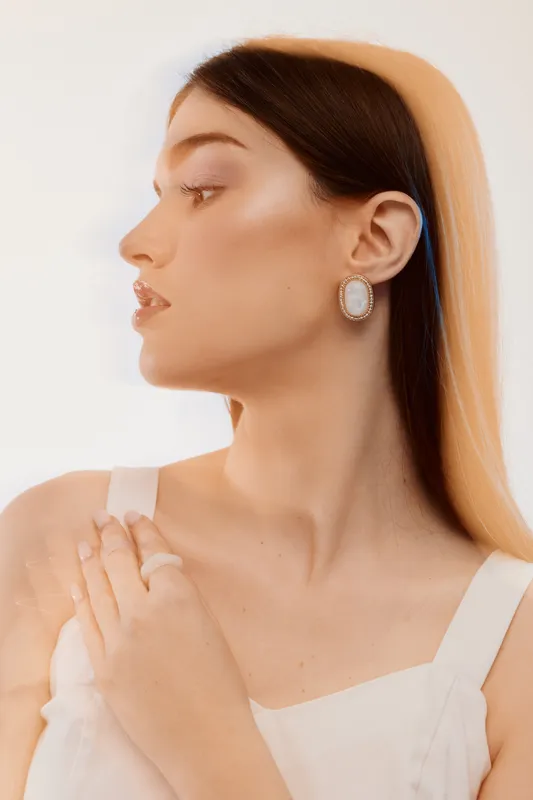 Elegant woman with smooth skin and long hair poses in white dress and statement earrings. Soft lighting highlights her beauty and fashion style in a minimalistic studio setting.