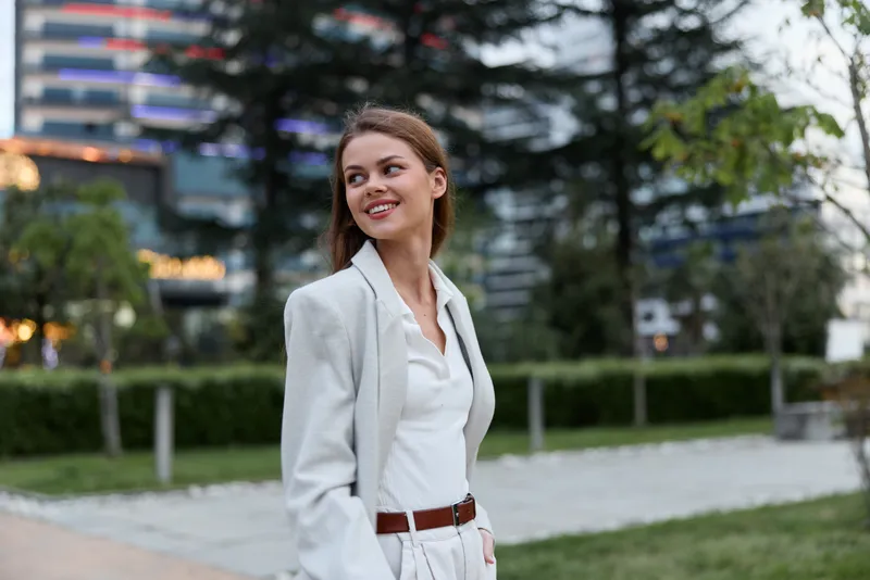 Young woman in white business suit smiling outdoors in urban park with modern buildings background, confident expression, natural light