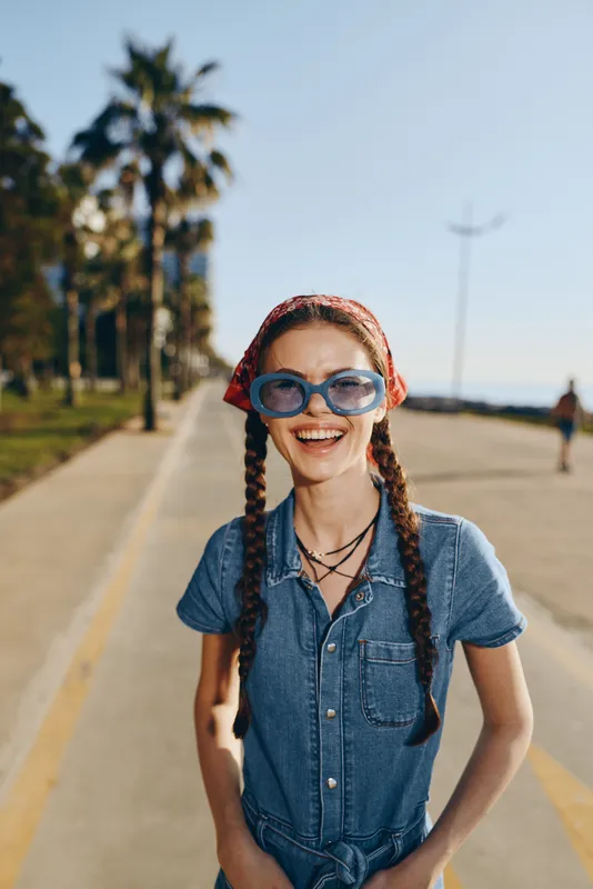 smiling woman wearing sunglasses and denim jumpsuit enjoying sunny day outdoors with palm trees on background and clear blue sky