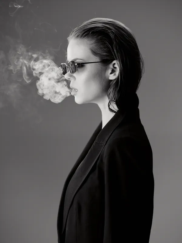 Black and white portrait of a stylish woman exhaling smoke wearing sunglasses and a dark blazer, moody and artistic atmosphere.