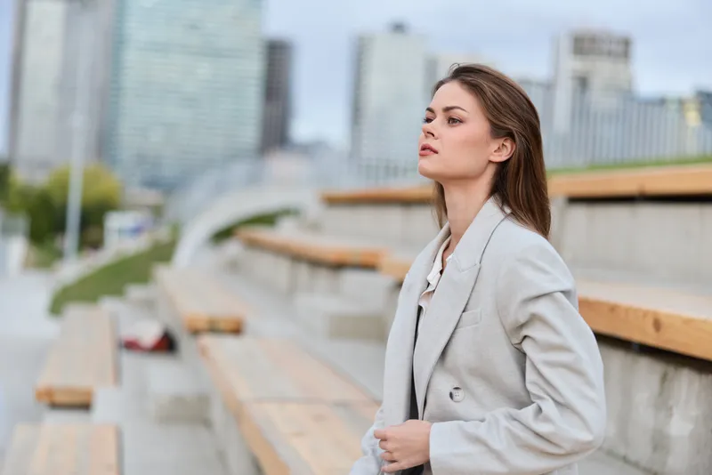Young confident woman in light gray coat sitting on outdoor benches with urban cityscape background in autumn season, thoughtful expression, business concept