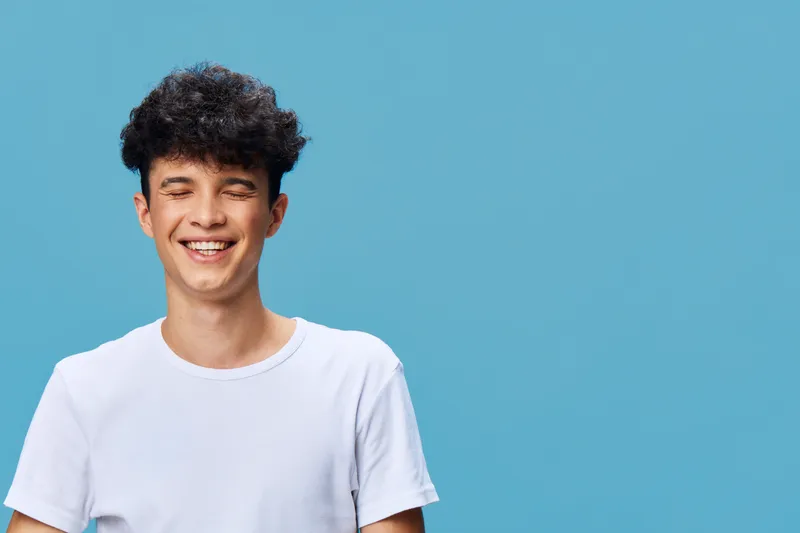 smiling man with curly hair wearing a white t-shirt against blue background, expressing happiness and youthfulness in casual style, bright and cheerful.