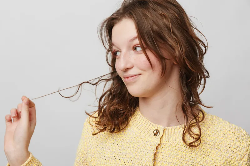 Young woman with curly brown hair wearing yellow knitted sweater plays with hair strands, looking sideways with a slight smile. Studio portrait on light grey background, casual mood.