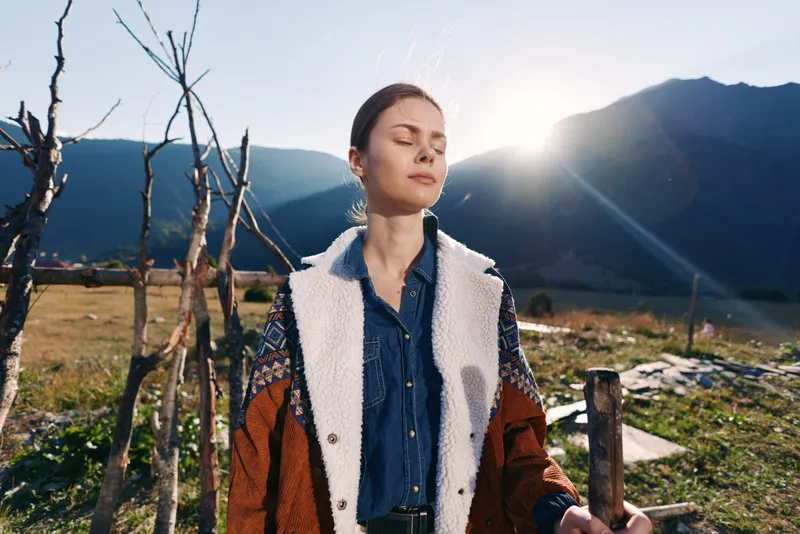 Woman portrait in mountains with sunlight and nature, peaceful outdoors scene. Closed eyes female in denim shirt and warm coat, relaxing by wooden fence in rural landscape at golden hour.