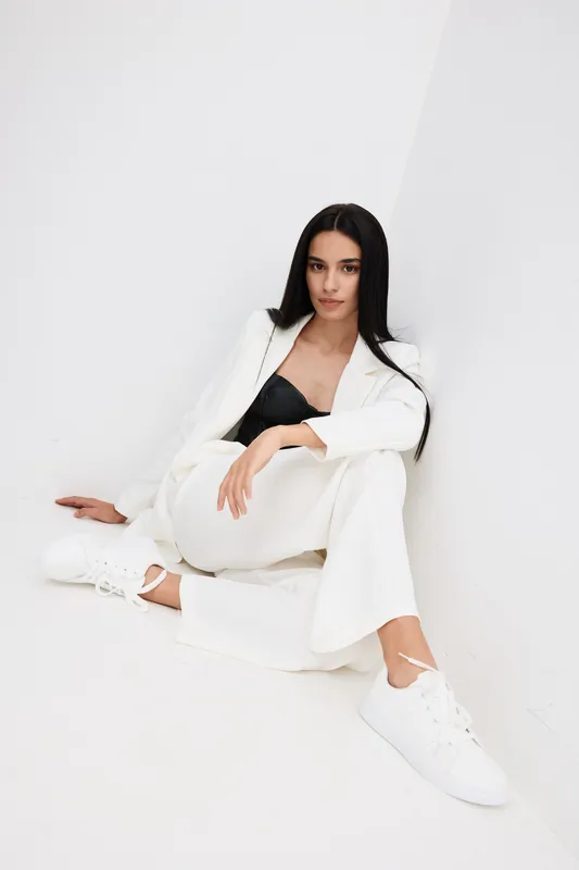 Stylish woman dressed in a white suit and black top sitting on the floor against a white wall with long dark hair and white sneakers, showcasing modern casual fashion.
