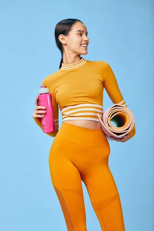 Woman fitness portrait: young woman in orange workout set holding a pink water bottle and rolled yoga mat, smiling and looking sideways against a clean blue studio background.
