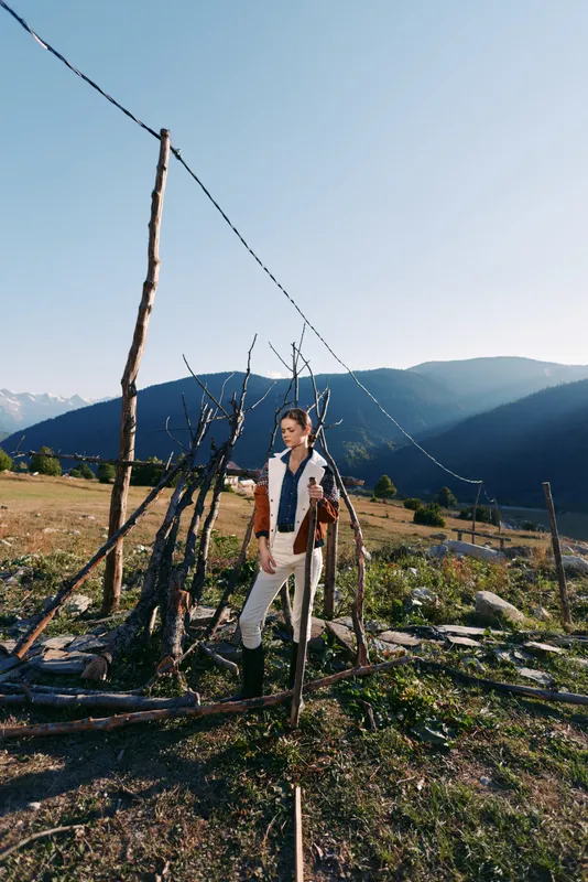 man mountains field fence standing portrait rural nature: young model in a rustic outdoor scene, standing by a makeshift fence in sunlight wearing casual vest and boots in scenic landscape