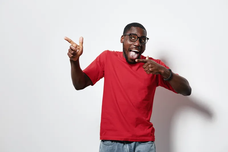 African American man with dark skin wearing glasses and red t shirt pointing fingers and smiling with tongue out on white background for fun and casual expression.