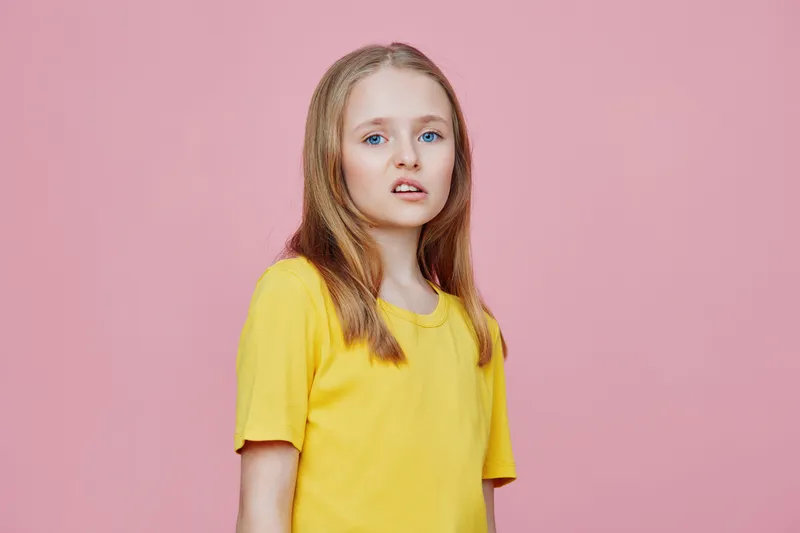 Portrait of a young girl with long hair wearing a yellow shirt against a pink background, expressing curiosity and innocence, ideal for children s themes