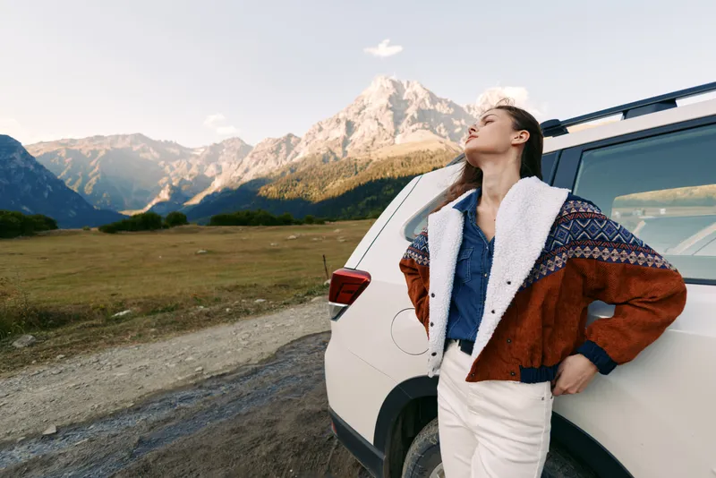 Woman leaning against white car at roadside with mountains background, travel roadtrip portrait in nature wearing warm patterned jacket, relaxed outdoors adventure and scenic view.