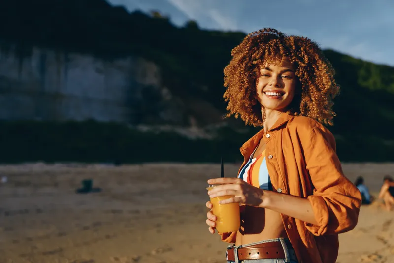 Smiling woman with curly hair on a beach, holding a drink cup, warm summer sunset light, outdoors nature travel vacation mood, relaxation and joy portrait.