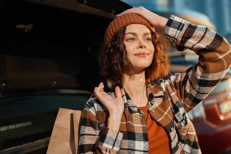 Woman portrait shopping by open car trunk, beanie and plaid jacket, candid smiling moment of urban lifestyle with golden hour glow, authenticity and mindful living vibe.