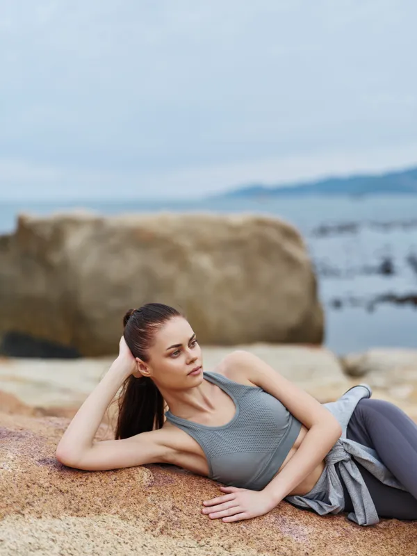 Young woman in activewear relaxing by the seaside, portraying a calm and confident demeanor in a natural outdoor setting, ideal for wellness and healthy lifestyle themes
