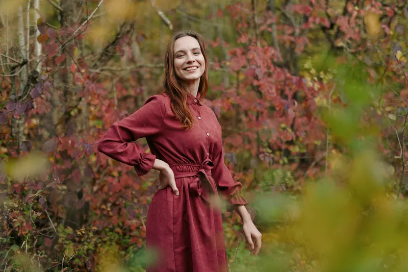 A genuine portrait of a smiling young woman in a red dress standing among autumn foliage, shot with natural light to evoke authenticity and believable everyday emotion and presence.