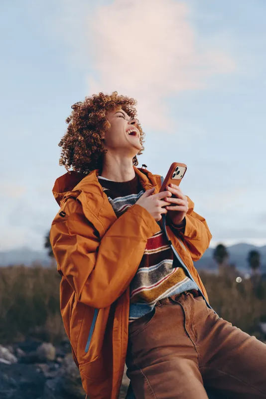 Woman lifestyle outdoors, curly hair, orange jacket, smiling using smartphone, holding phone near face, nature landscape and cloudy sky, travel mood in countryside during daytime today