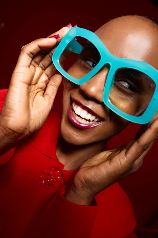 Close-up portrait of a smiling black woman with dark skin wearing oversized turquoise glasses and a red top. Bright red background creates a vibrant and joyful fashion concept.