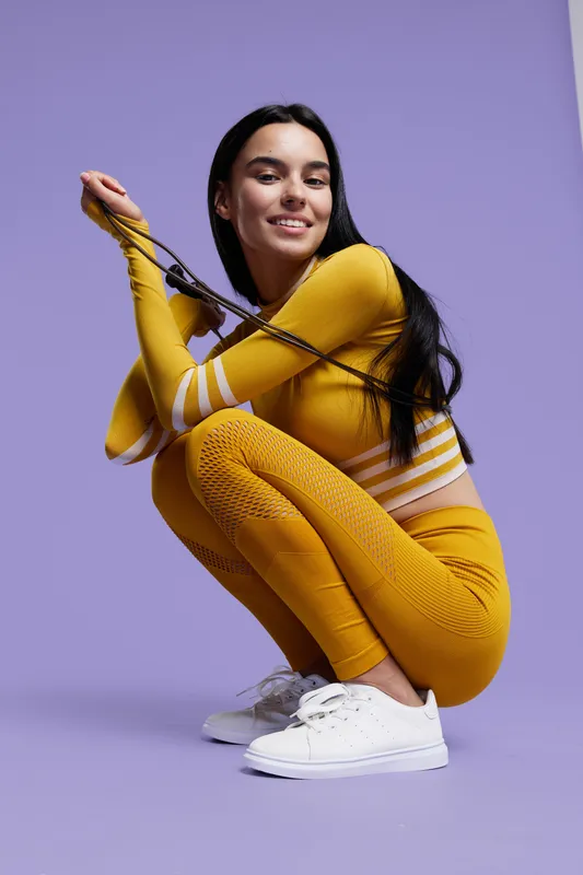 Woman sport fitness wearing yellow activewear squatting while smiling confidently with jump rope on purple background, showing energy and healthy lifestyle.