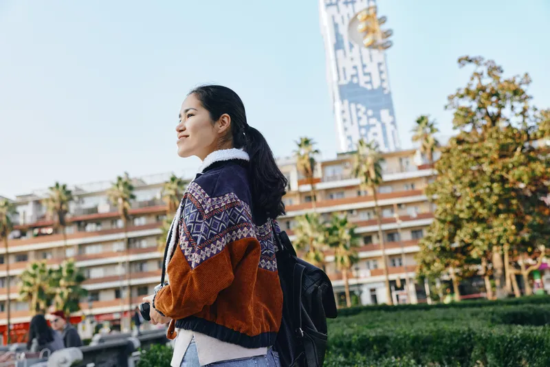 Woman with backpack wearing cozy sweater walks outdoors along a sunny street with palm trees and modern buildings, casual fashion, daily urban errands and fresh air