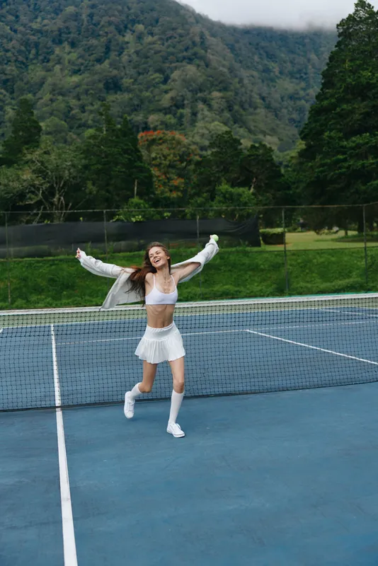 Young athletic woman joyfully celebrating after winning a tennis match on an outdoor court surrounded by greenery and mountains, wearing a stylish white sports outfit
