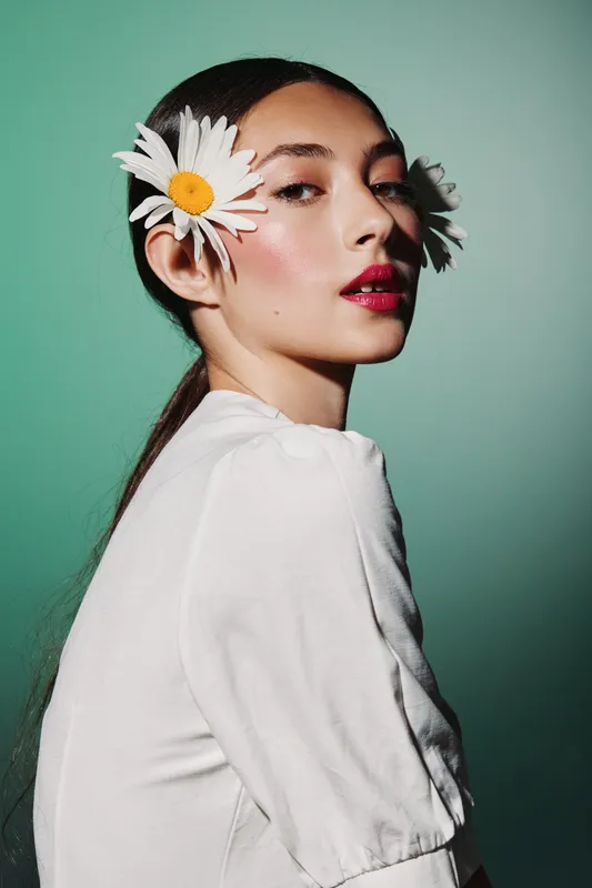 Young woman with fresh makeup and glossy lips styled with white daisies in her hair. Studio shot with green background. Concept of beauty fashion with natural floral elements.