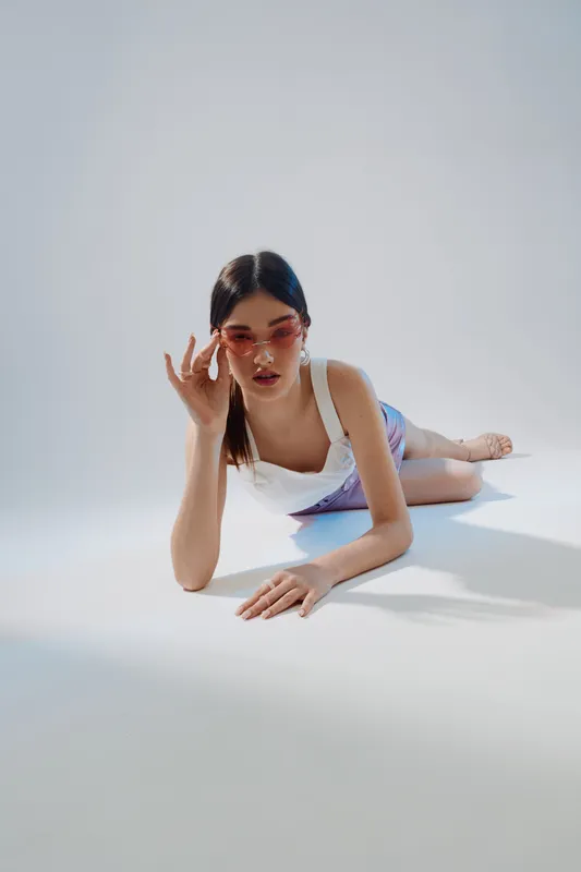 Young woman in white outfit and pink sunglasses lies on the floor in a studio with soft lighting. Concept of beauty, fashion, and modern style for editorial or advertising.