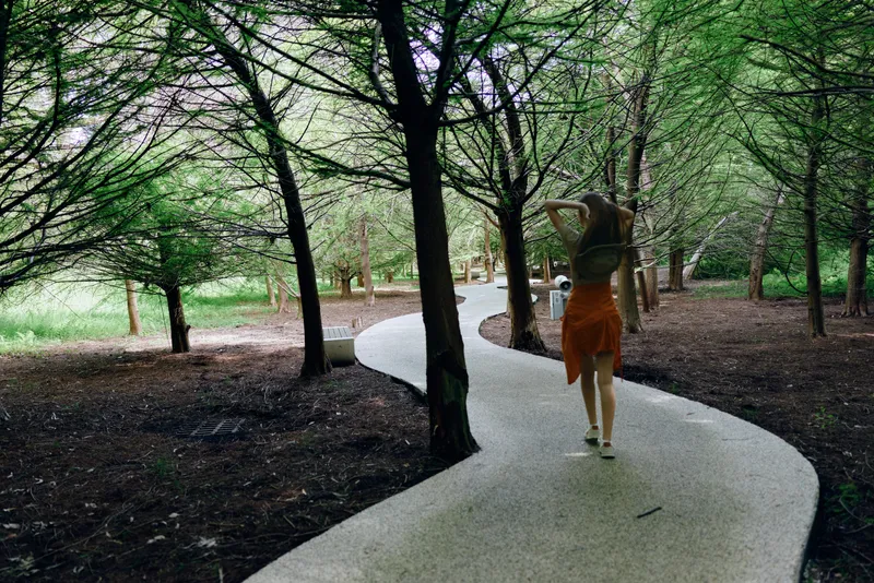 Woman walking on a curved path through trees in a shaded park, carrying a backpack and wearing a summer dress. Forest trail scene for outdoor leisure and peaceful nature stroll.