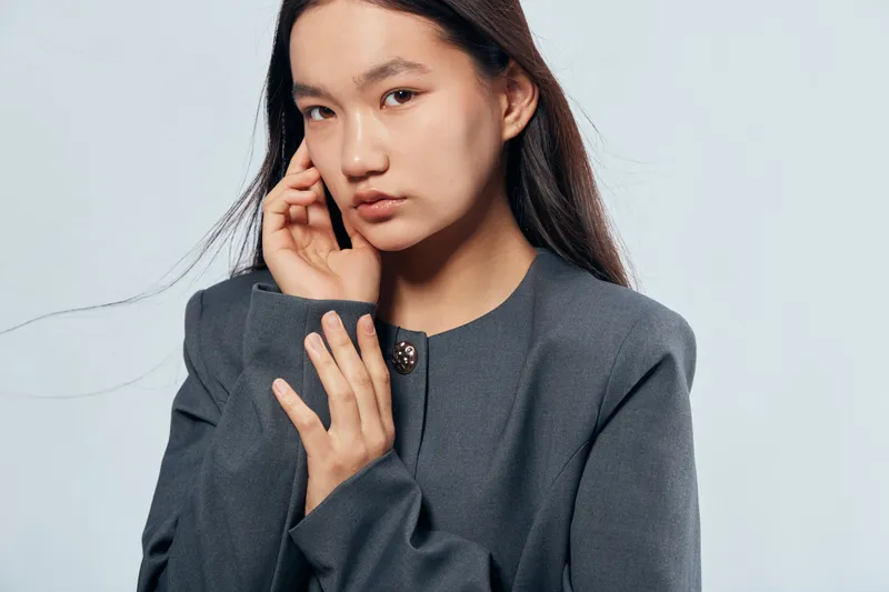 Young woman with long dark hair posing in gray jacket on plain background looking pensively at camera with hand near face in studio portrait with soft lighting and neutral expression.