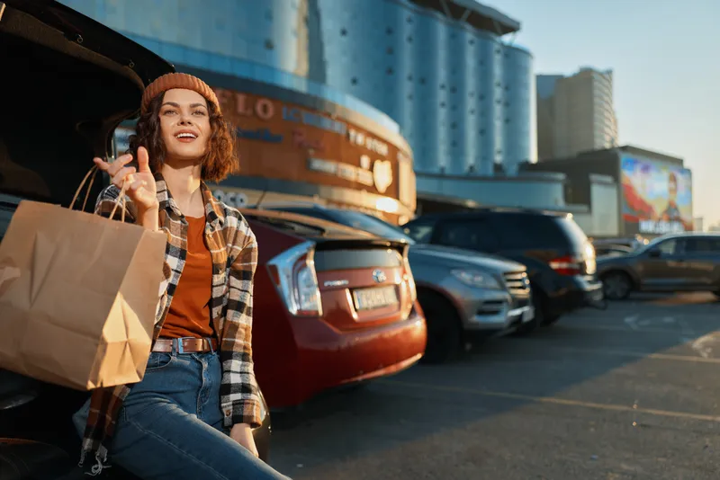 Woman shopping in urban parking by car, smiling with paper bag and beanie, casual style during golden hour glow, candid lifestyle authenticity and emotional storytelling of mindful living.