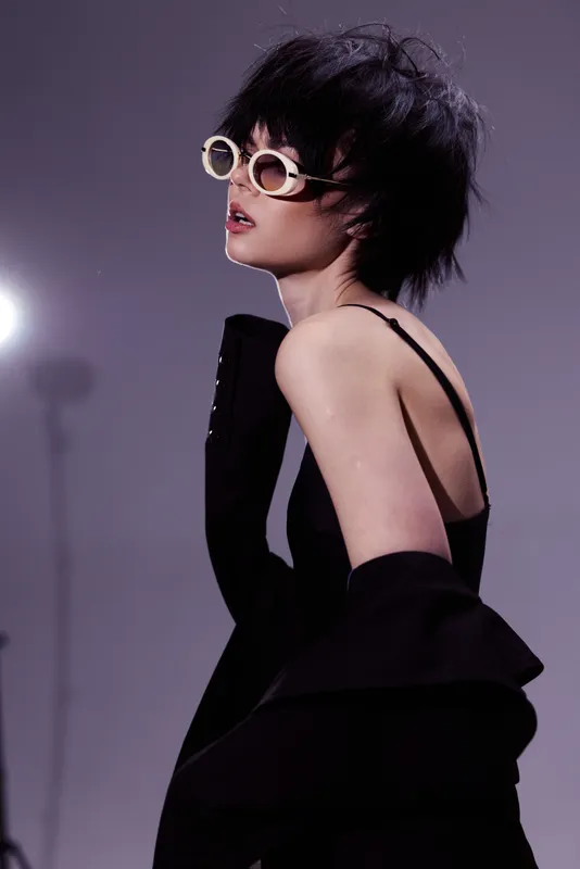 Stylish woman wearing sunglasses and black dress in a moody studio with dramatic lighting and modern fashion vibe.