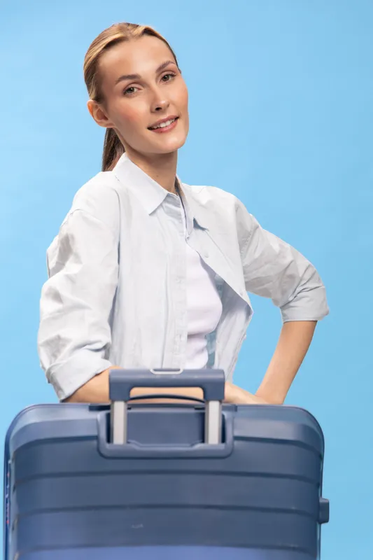Young woman with suitcase stands against blue background, smiling confidently. Casual outfit with white shirt and light makeup. Concept for travel, vacation, and lifestyle.