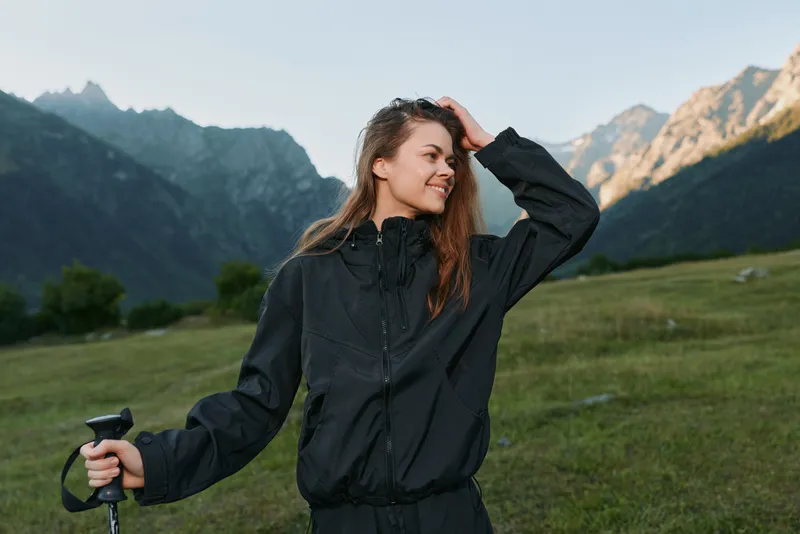 A cheerful hiker stands in a grassy alpine field, surrounded by towering mountains. She smiles softly, griping a trekking pole, exuding confidence and a sense of adventure in the clean, open air.