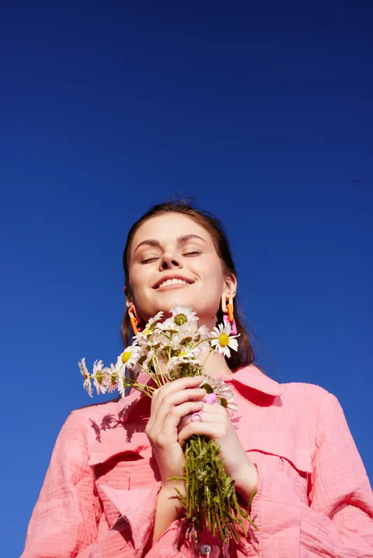 Young woman with cheerful expression holding wildflowers against a clear blue sky, wearing a pink shirt, symbolizing joy and connection with nature