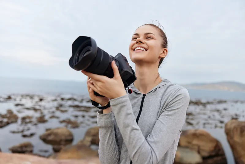 Young woman with camera smiling joyfully on the rocky beach at sunset, capturing memories in nature Photography passion and adventure concept