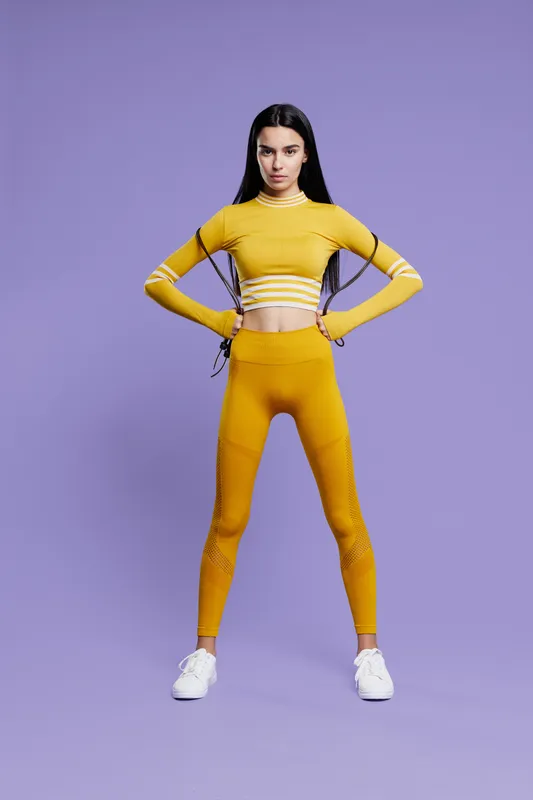 woman sport fitness wearing yellow activewear stands confidently with hands on hips against violet background, showcasing strength, focus, and stylish workout attire.