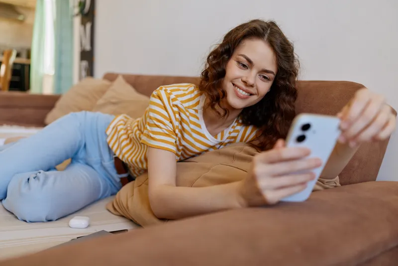 A relaxed woman lounges on a sofa at home, taking a casual selfie with a smartphone. A cozy living room setting communicates everyday comfort and modern home life.