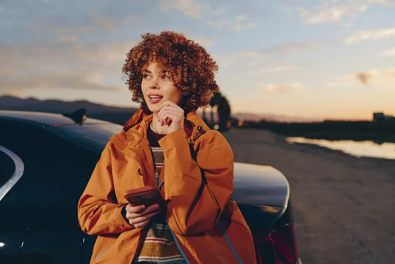 Woman lifestyle outdoors at sunset beside a parked car, curly hair and orange jacket, holding a smartphone, smiling on a quiet road with mountains in distance, travel mood, relaxed