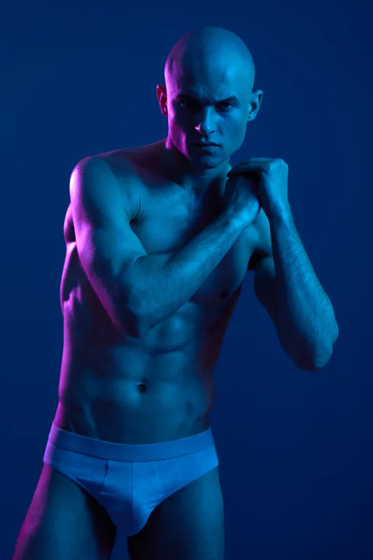 Athletic man posing under dramatic lighting with blue and purple tones, showcasing muscle definition and strong expression, emphasizing strength and confidence.