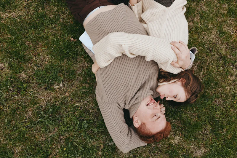 Young couple lying on grass in cozy sweaters share a joyful moment together outdoors in a relaxed lifestyle scene during autumn.