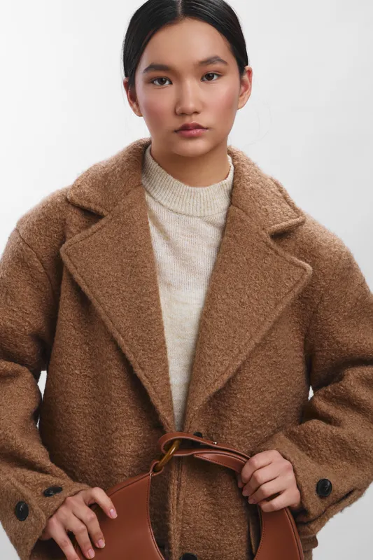 Asian woman in a textured brown coat stands against a plain backdrop, exuding poised confidence and modern style, holding a leather bag strap, presenting a polished image suitable for fashion and