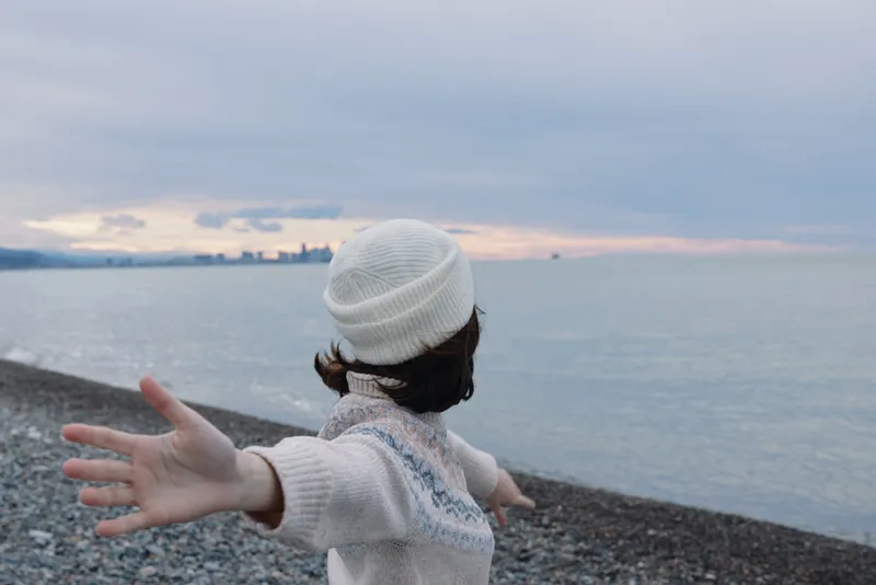 Child with a white knit hat on a pebble beach reaching toward the sea at sunset, a moment of open wonder by the calm water and distant horizon.