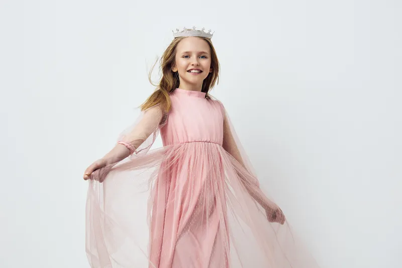 Young girl in a pink dress and crown dancing joyfully against a light backdrop, embodying innocence and playfulness for children s fashion or celebrations