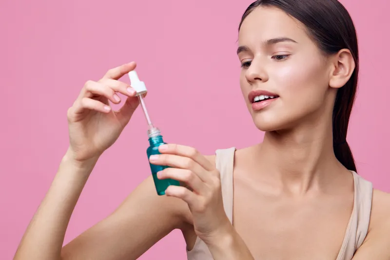 Young woman applying serum with dropper, showcasing a healthy skincare routine against a vibrant pink background, emphasizing beauty and self care