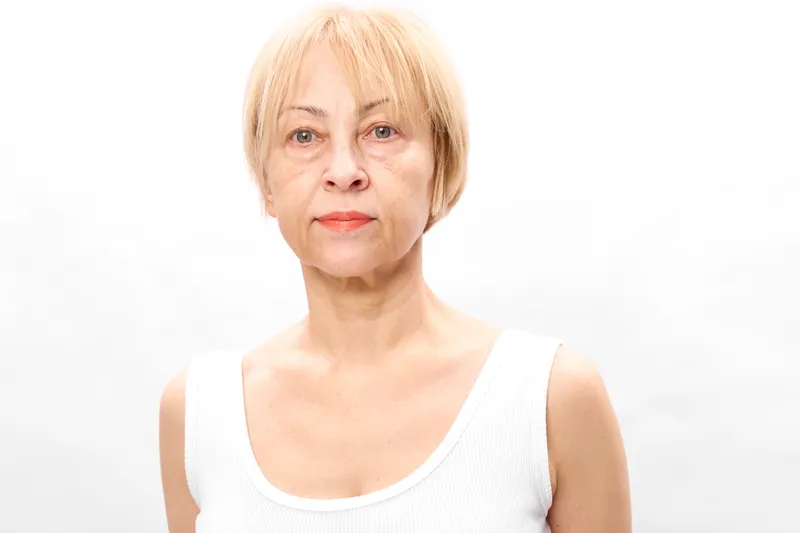 Senior woman in studio portrait, blonde short hair, neutral expression, looking at camera, plain white background, soft light, calm mood, minimal composition, soft shadows, close up